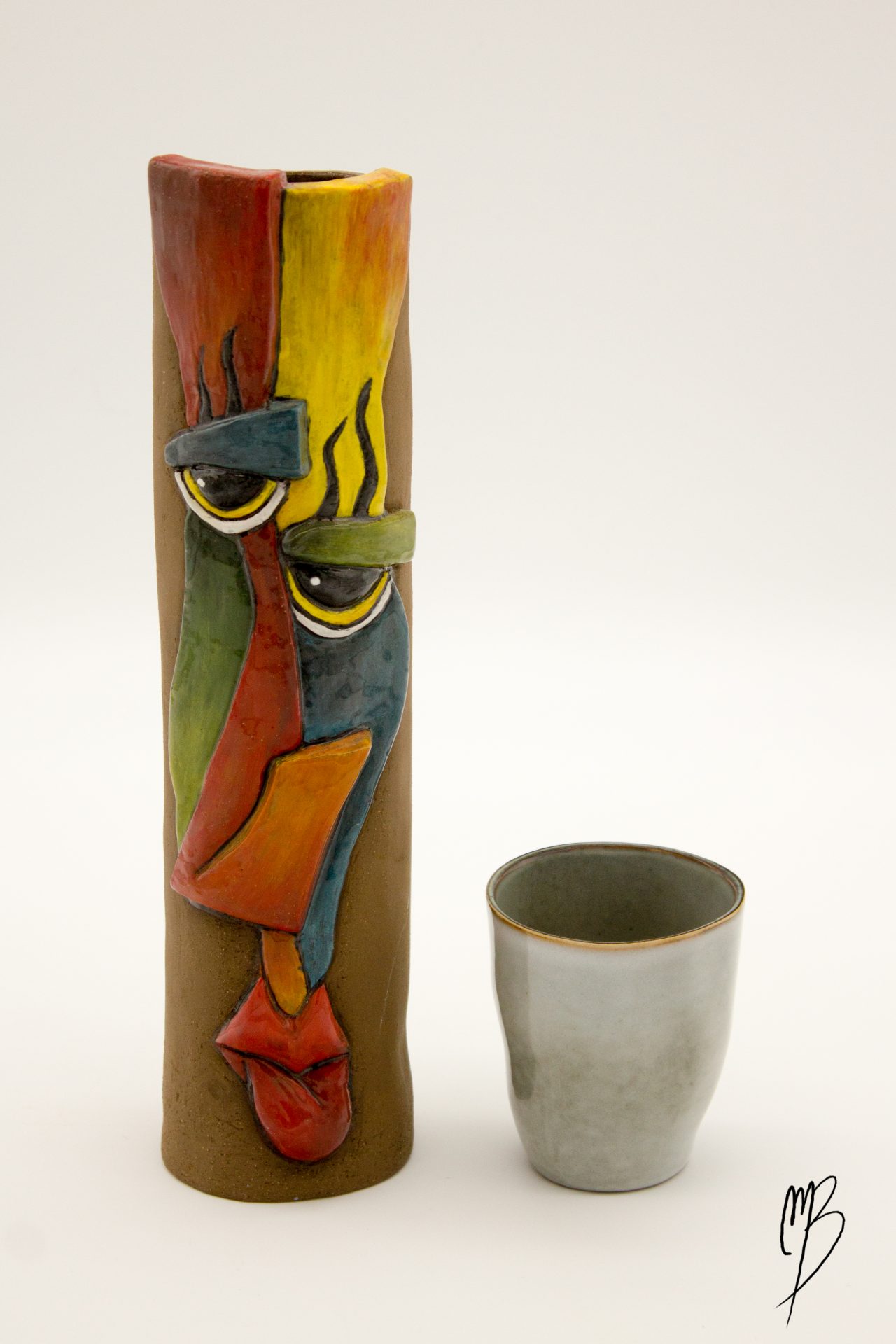 modelages sculptures d'un vase haut et fin orné d'un visage abstrait rouge jaune vert et bleu en terre émaillé, accompagné d'une tasse pour refléter la taille du vase