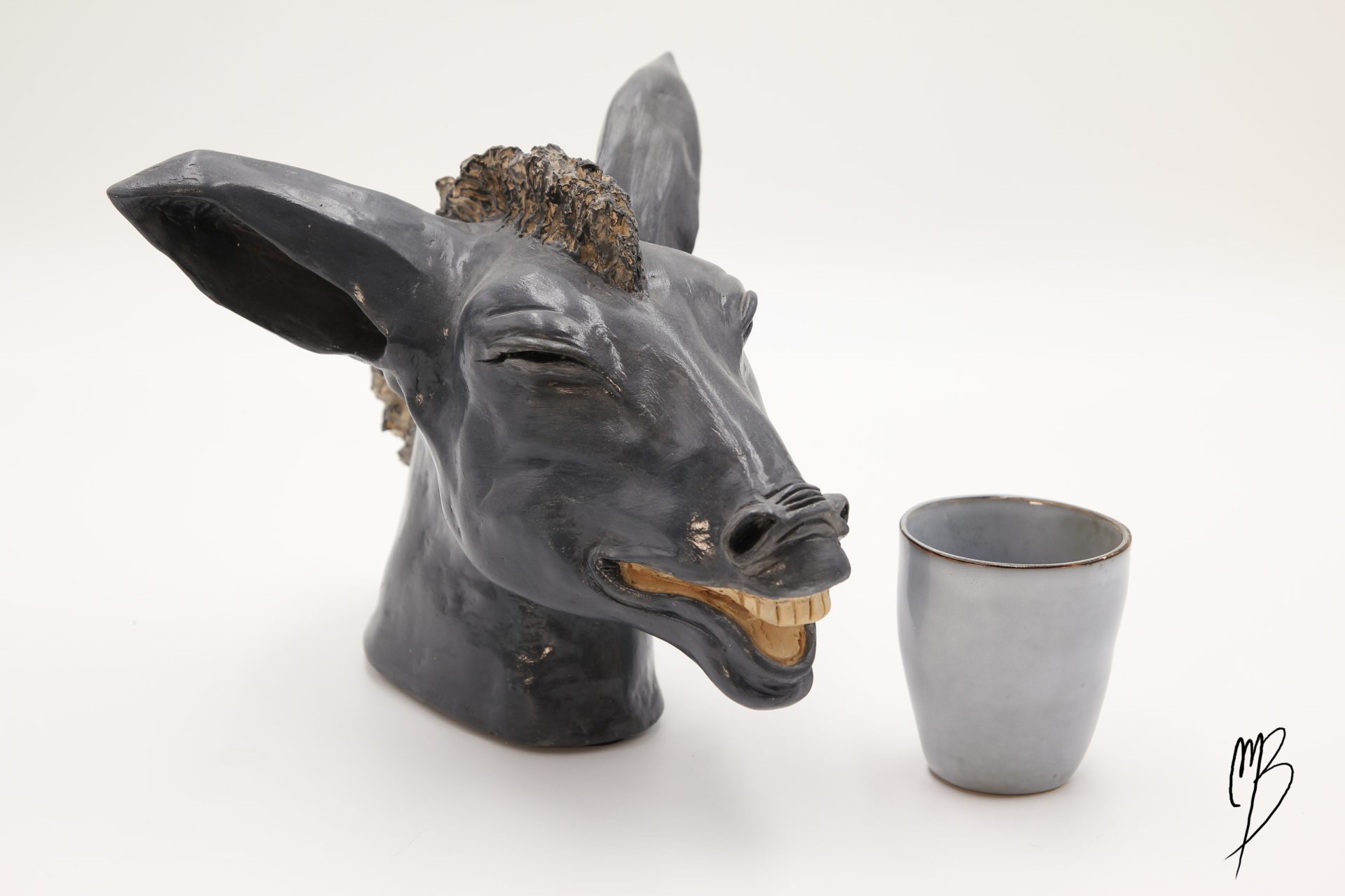 Animaux. Tête d'âne rieur et moquer en terre cuite recouverte d'une peinture acrylique noire vernie. Vue comparative à une tasse pour la taille.