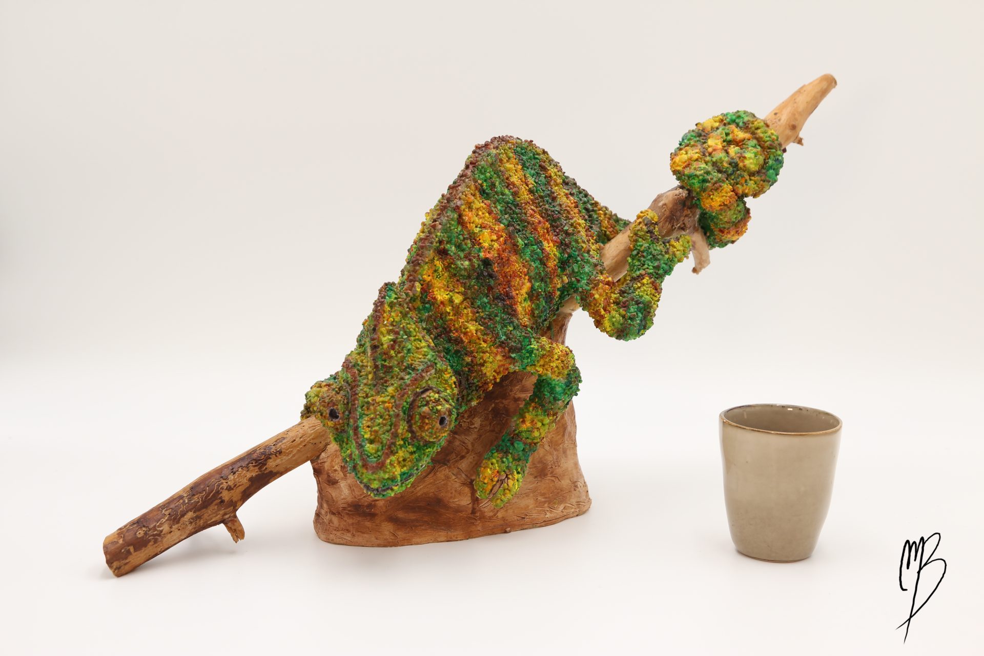 Animaux. Caméléon sur sa branche en terre cuite recouvert de sable et de peinture acrylique vernie. Vue comparative à une tasse pour la taille