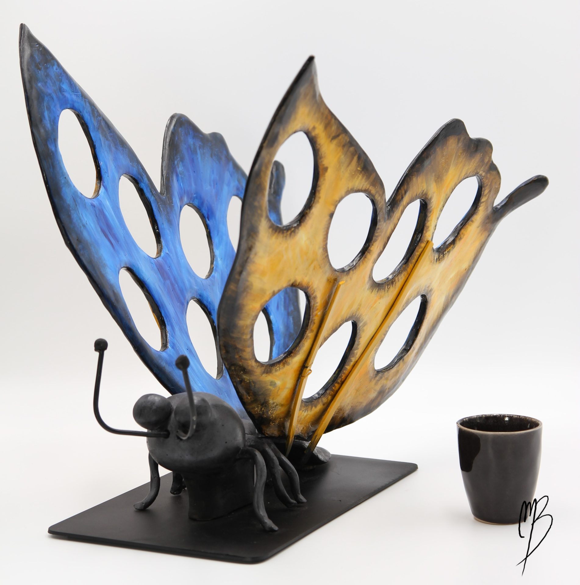 Animaux. Morpho Bleu aux ailes déployées jaunes et bleues sur un socle en métal en terre cuite recouverts de peinture acrylique vernie. Vue comparative à une tasse pour la taille
