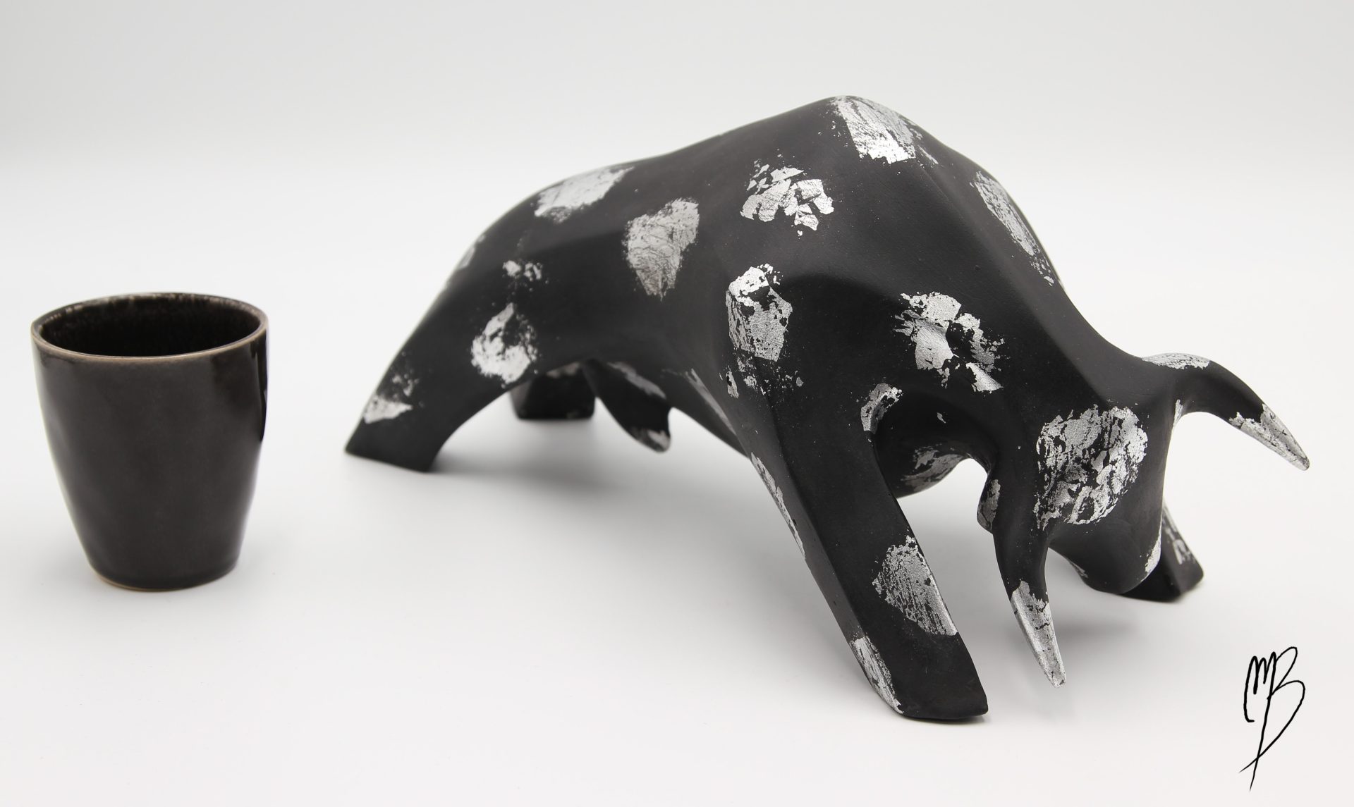 Animaux. Taureau noir en terre cuite recouvert de peinture acrylique et de feuilles d'argent. Vue comparative à une tasse pour la taille.