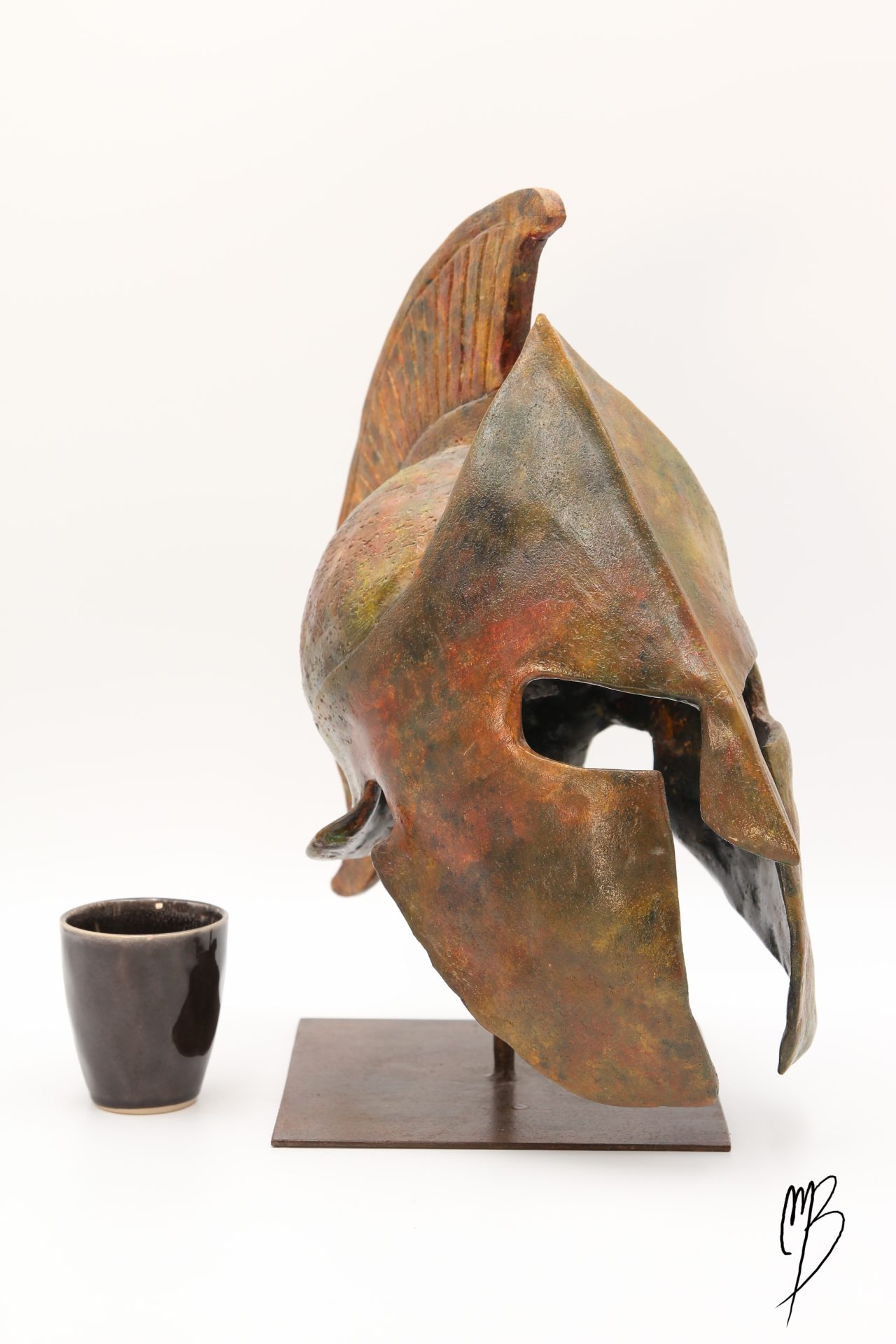 Casque Spartiate posé sur un support métallique en Terre cuite patinée avec des peintures acryliques imitant des bronzes anciens. Vue comparative à une tasse pour la taille.
