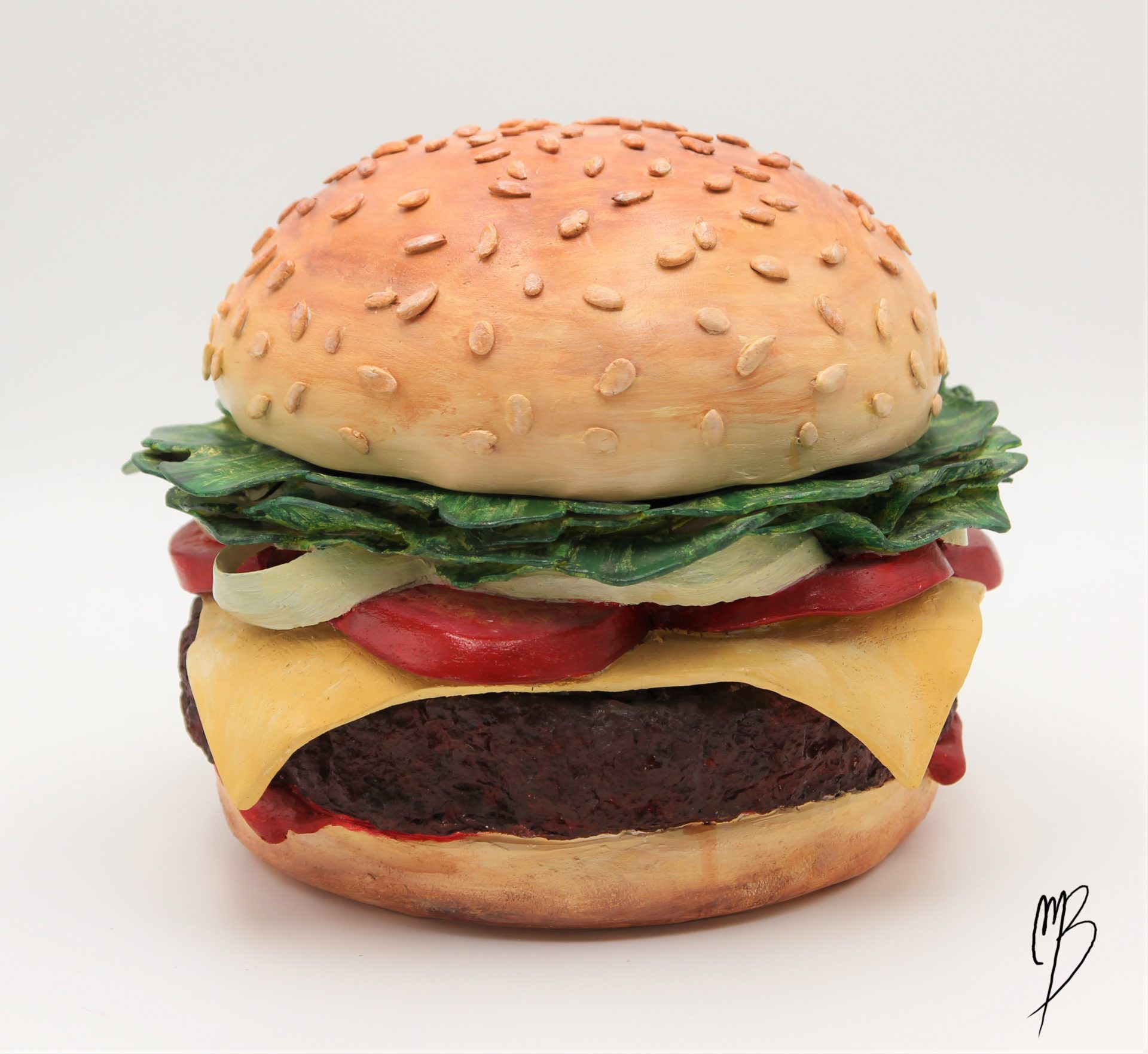 Sculpture en terre cuite patinée avec des peintures acryliques rouges vertes jaunes marrons. Elle représente un hamburger composée de deux tranches de pain, un steak haché, des tomates en tranche, du fromage fondu et de la salade. Vue générale de face