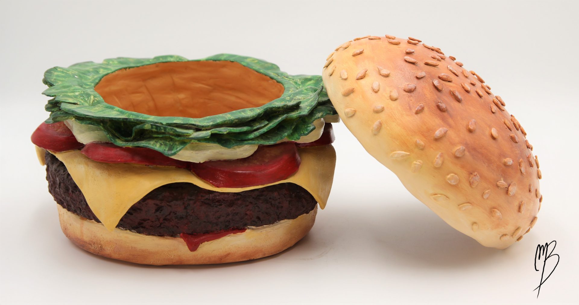 Terre cuite patinée avec des peintures acryliques rouges vertes jaunes marrons. Elle représente un hamburger composée de deux tranches de pain, un steak haché, des tomates en tranche, du fromage fondu et de la salade. Vue avec le pain du dessus déposé sur le côté