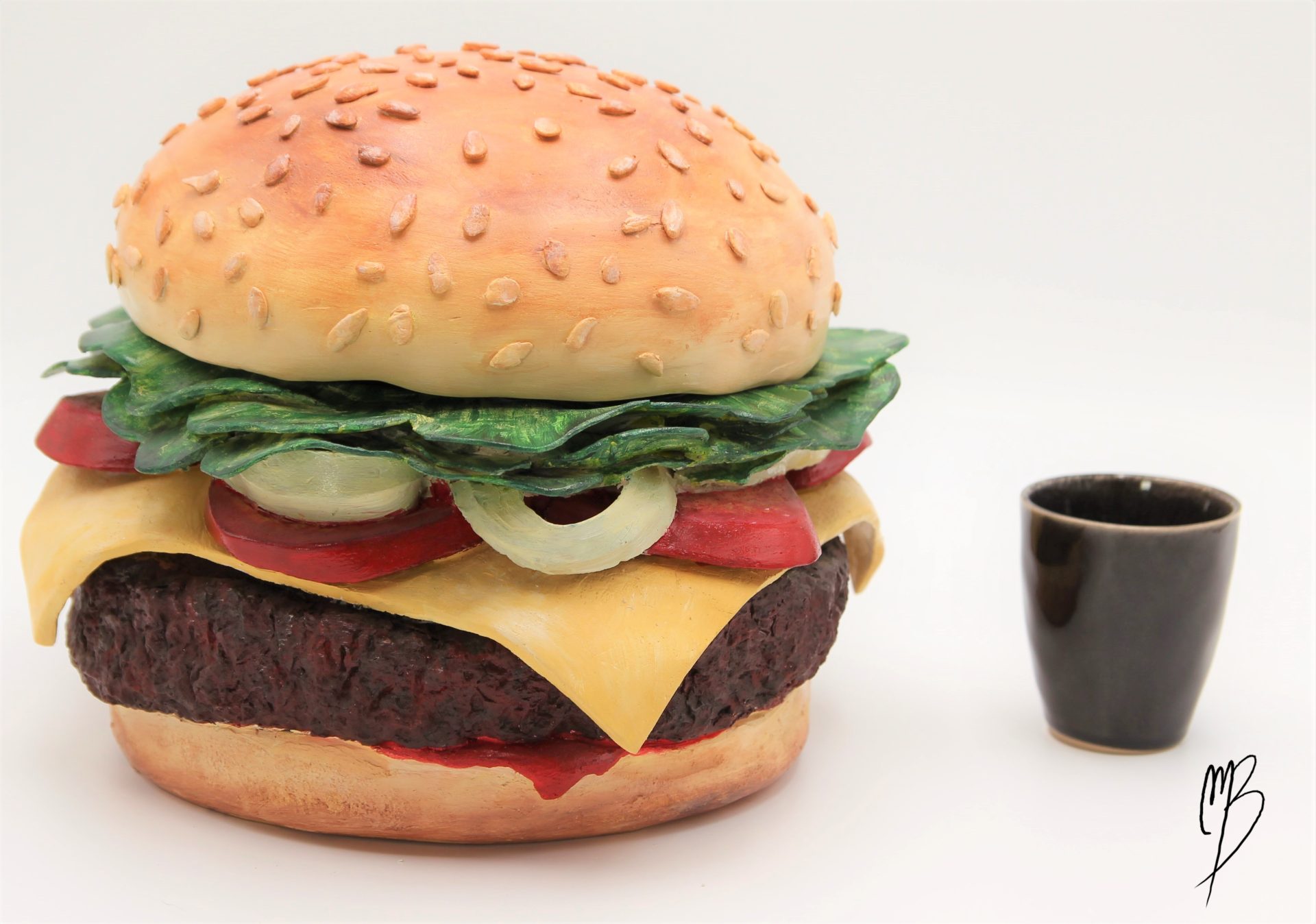 Terre cuite patinée avec des peintures acryliques rouges vertes jaunes marrons. Elle représente un hamburger composée de deux tranches de pain, un steak haché, des tomates en tranche, du fromage fondu et de la salade. Vue comparative à une tasse précisant la taille.