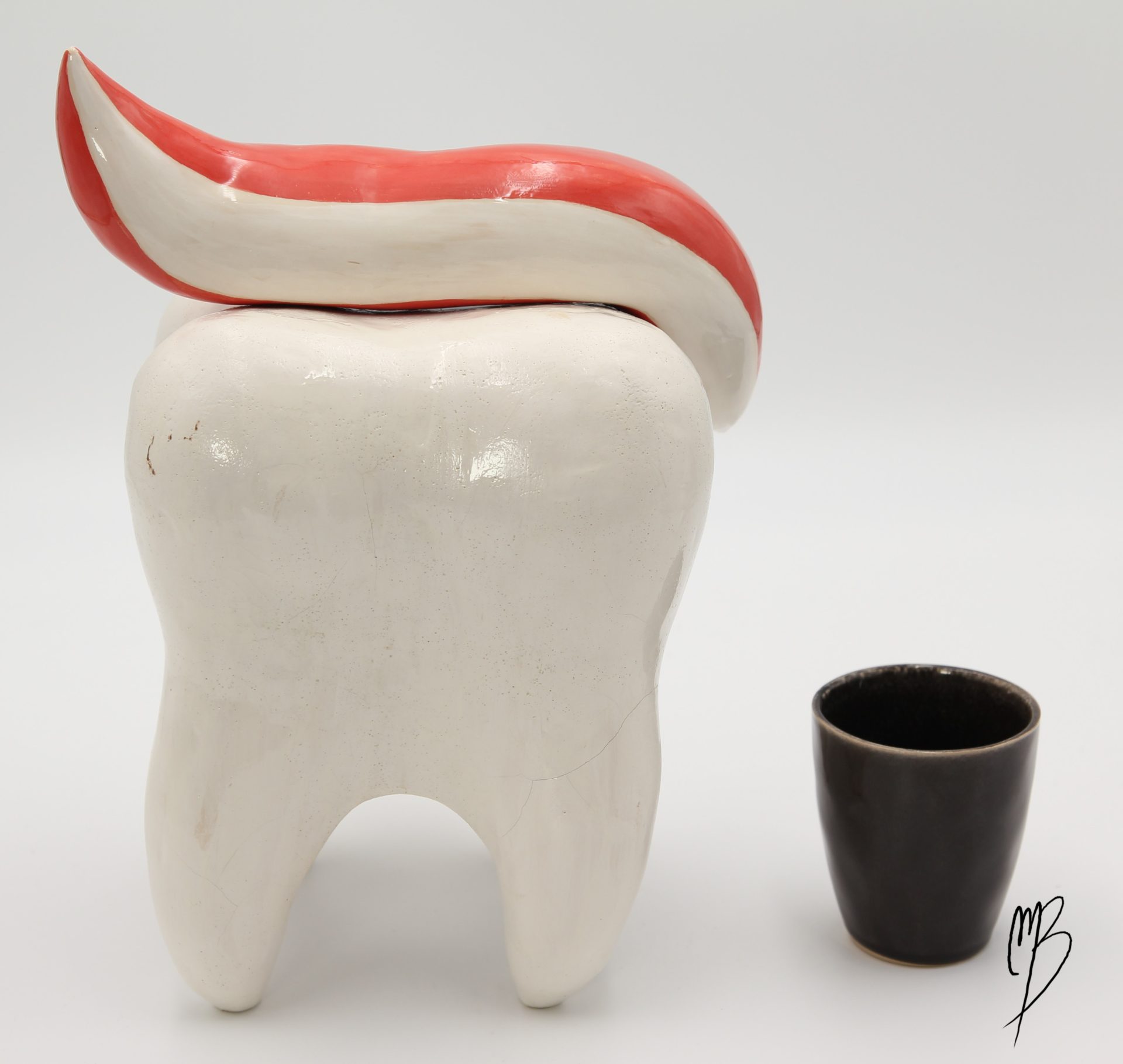 Dent blanche émaillée vernie reposant sur ses racines recouverte de dentifrice rouge et blanc. Vue comparative à une tasse pour la taille.