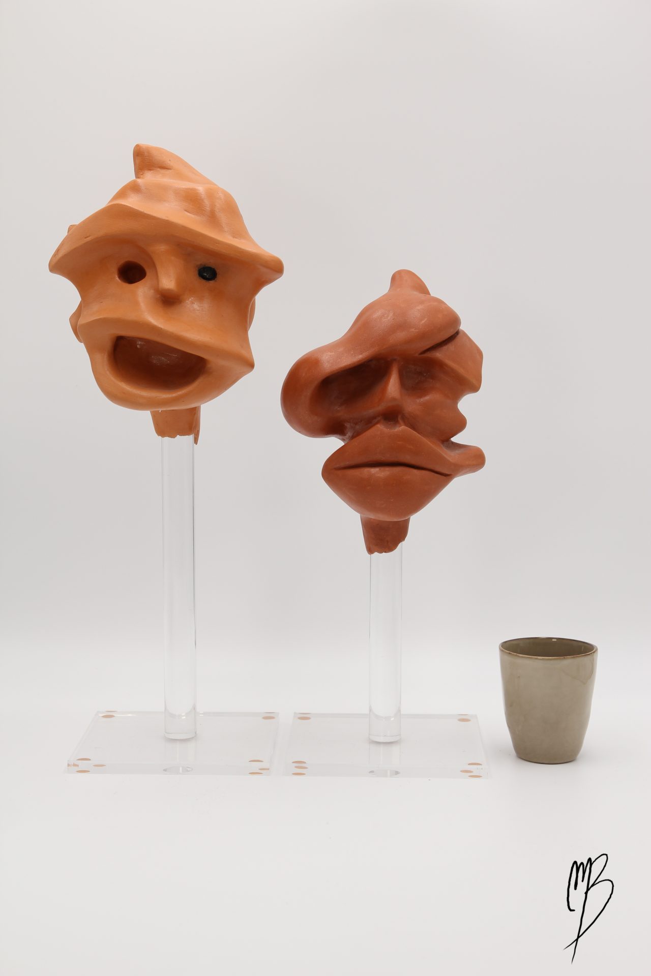 Deux prises d'escalade représentant deux têtes de primate en terre cuite vernie reposant sur deux socle transparent en plexiglass. Vue comparative à une tasse pour la taille
